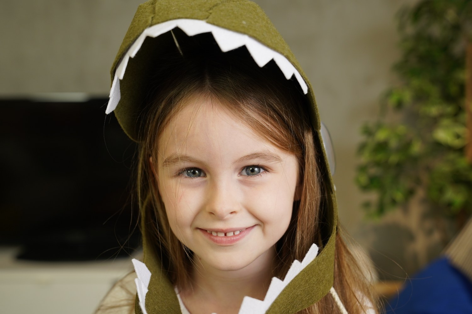 A close-up portrait of a smiling young girl with long brown hair and blue eyes, wearing a green felt dinosaur hood with white jagged teeth framing her face.